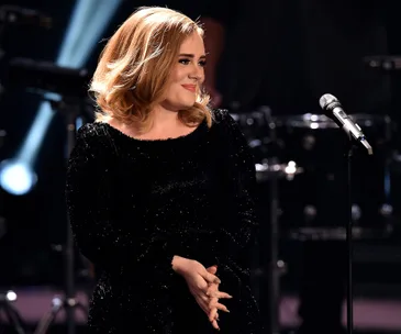 Adele with a new hairstyle, wearing a black dress and smiling on stage near a microphone.