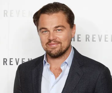 A man in a suit at an event with "The Revenant" in the background.