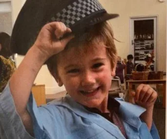A young child smiling, wearing a uniform with a checkered hat indoors.