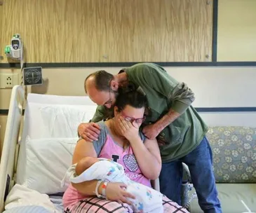 A woman holding a newborn and crying on a hospital bed, comforted by a man hugging her from behind.
