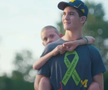 Young man carrying a boy on his back, wearing a cap and a T-shirt with a cerebral palsy awareness ribbon.