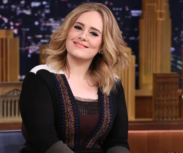 A person with shoulder-length hair, smiling and sitting on a talk show set with a cityscape backdrop.