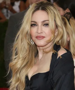 A person with long blonde hair and a black outfit, attending an event, standing in front of a blurred background.