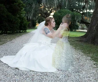 Bride in a white dress kneels on a path, touching a ghostly image of a young girl, surrounded by greenery.