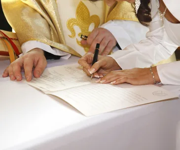 Bride signing a marriage document, overseen by a person in ceremonial attire.