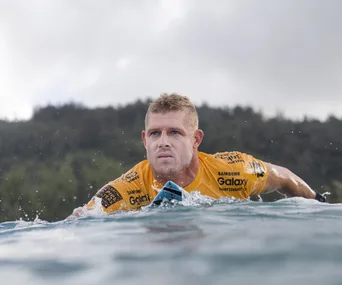 Surfer in a yellow jersey paddles on a surfboard with a focused expression amid ocean waves.
