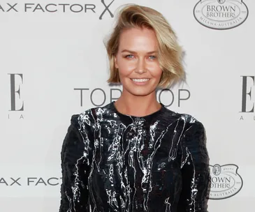 A person smiling in a shiny black outfit on a red carpet at a fashion event backdrop.