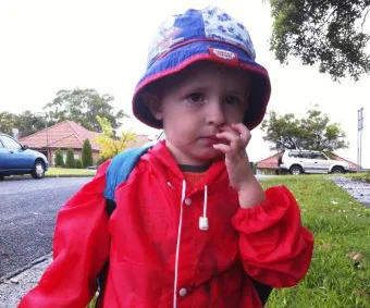 Young child in a red raincoat and hat, standing outdoors, touching face with trees and parked cars in background.