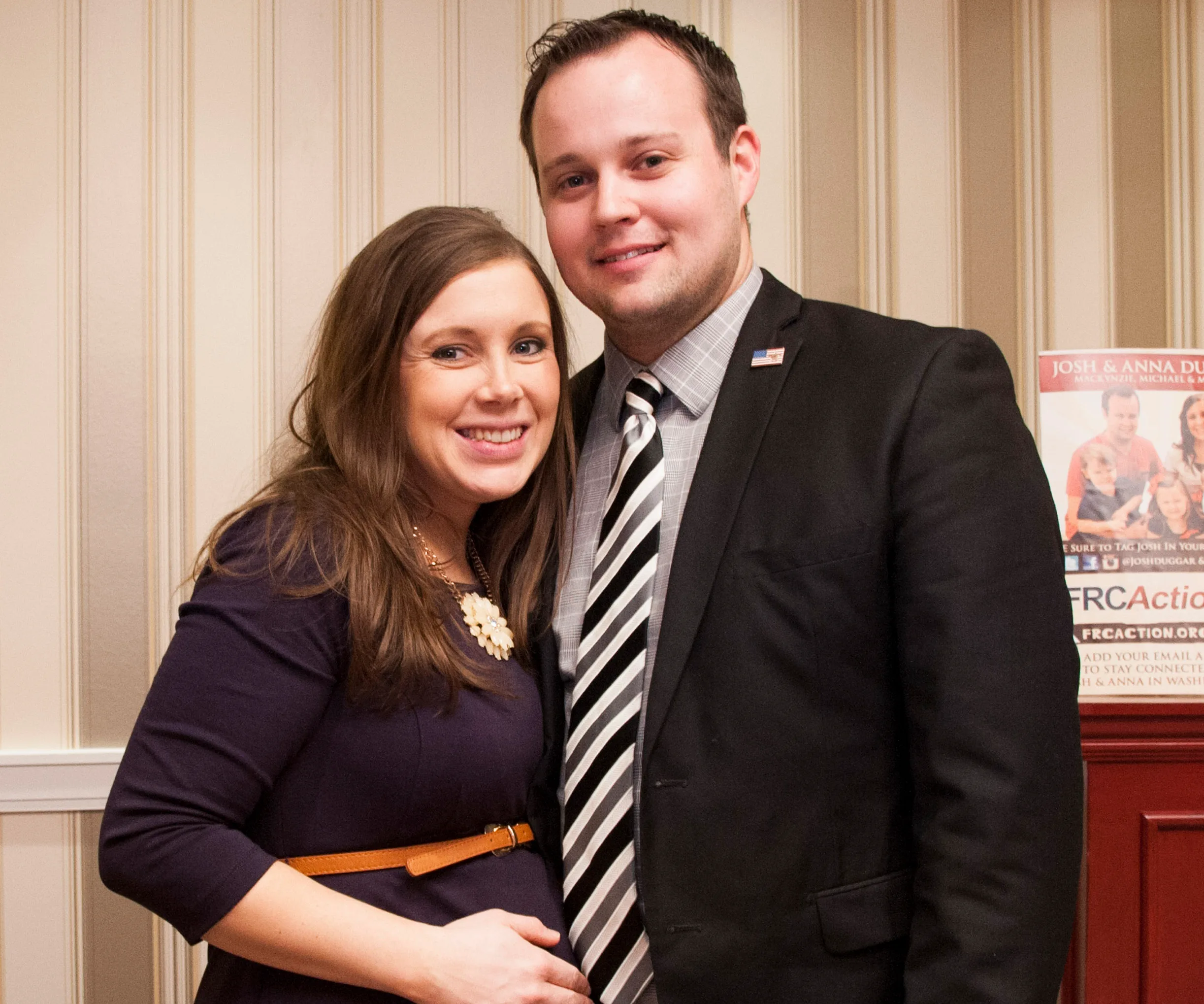 A couple smiling together in front of a striped wall, with the woman holding her pregnant belly.