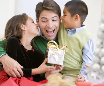 Father joyfully receiving a gift from his two children, with a Christmas tree nearby.