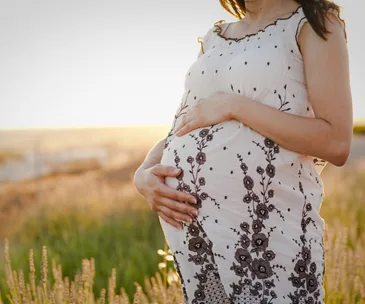 Pregnant woman in a floral dress holding her belly in a sunny outdoor setting.