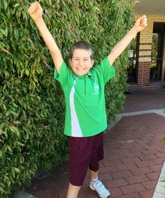 Young boy in green shirt raises arms victoriously, smiling outdoors against leafy background.