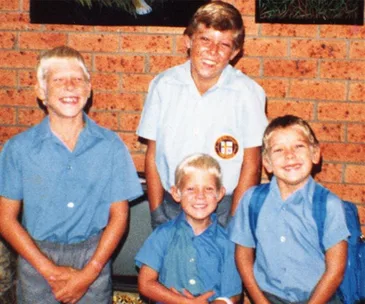 Four young boys in school uniforms smiling in front of a brick wall.