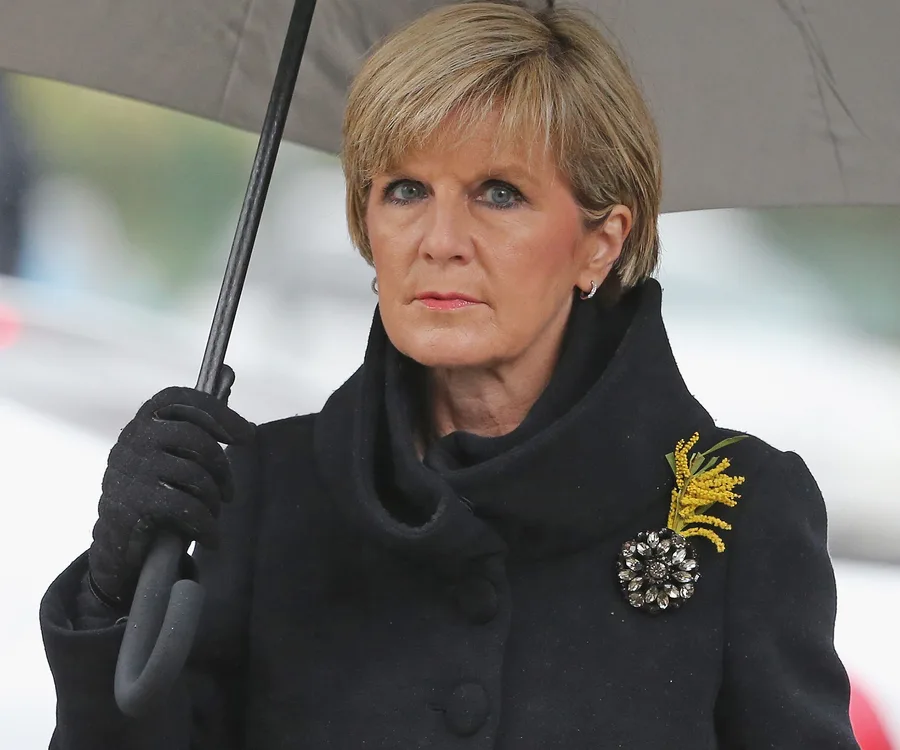 A woman in a black coat holds an umbrella, wearing a brooch and a serious expression, standing outdoors.