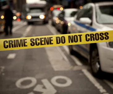Yellow "Crime Scene Do Not Cross" tape blocks a street with blurred cars in the background.