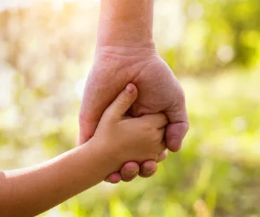 An adult and child holding hands against a blurred, sunny background.