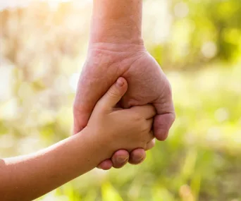 An adult and child holding hands against a blurred, sunny background.