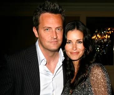 A man and a woman smiling and posing together at a formal event with chandeliers in the background.