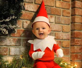 Baby dressed as Elf on the Shelf, wearing a red outfit and elf hat, sits smiling against a brick wall with greenery.