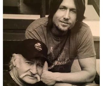 "Man with mustache wearing cap sitting beside man in t-shirt on steps, black and white photo."