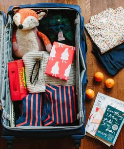 Open suitcase with folded clothes, a striped stuffed toy, red gift boxes, and oranges beside magazines on the wooden floor.