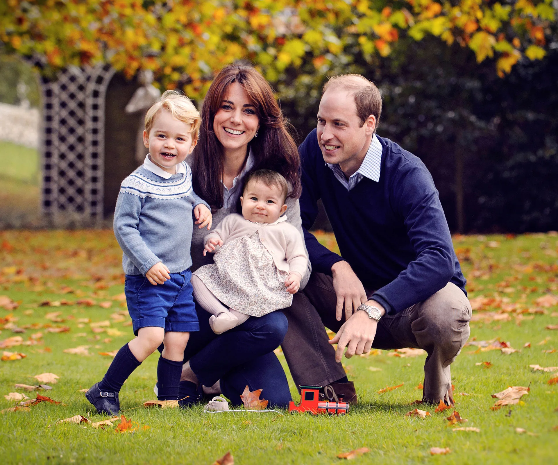 A family of four, two adults and two children, pose on autumn grass with scattered leaves.