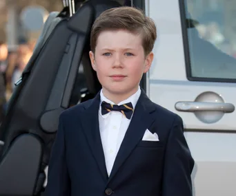 A young boy in a suit and bow tie stands in front of a vehicle.