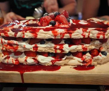 Layered meringue cake with strawberries, blueberries, cream, and red fruit sauce on a wooden board.