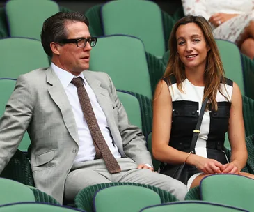 Man in a gray suit and woman in black and white dress sit together in green stadium seats.