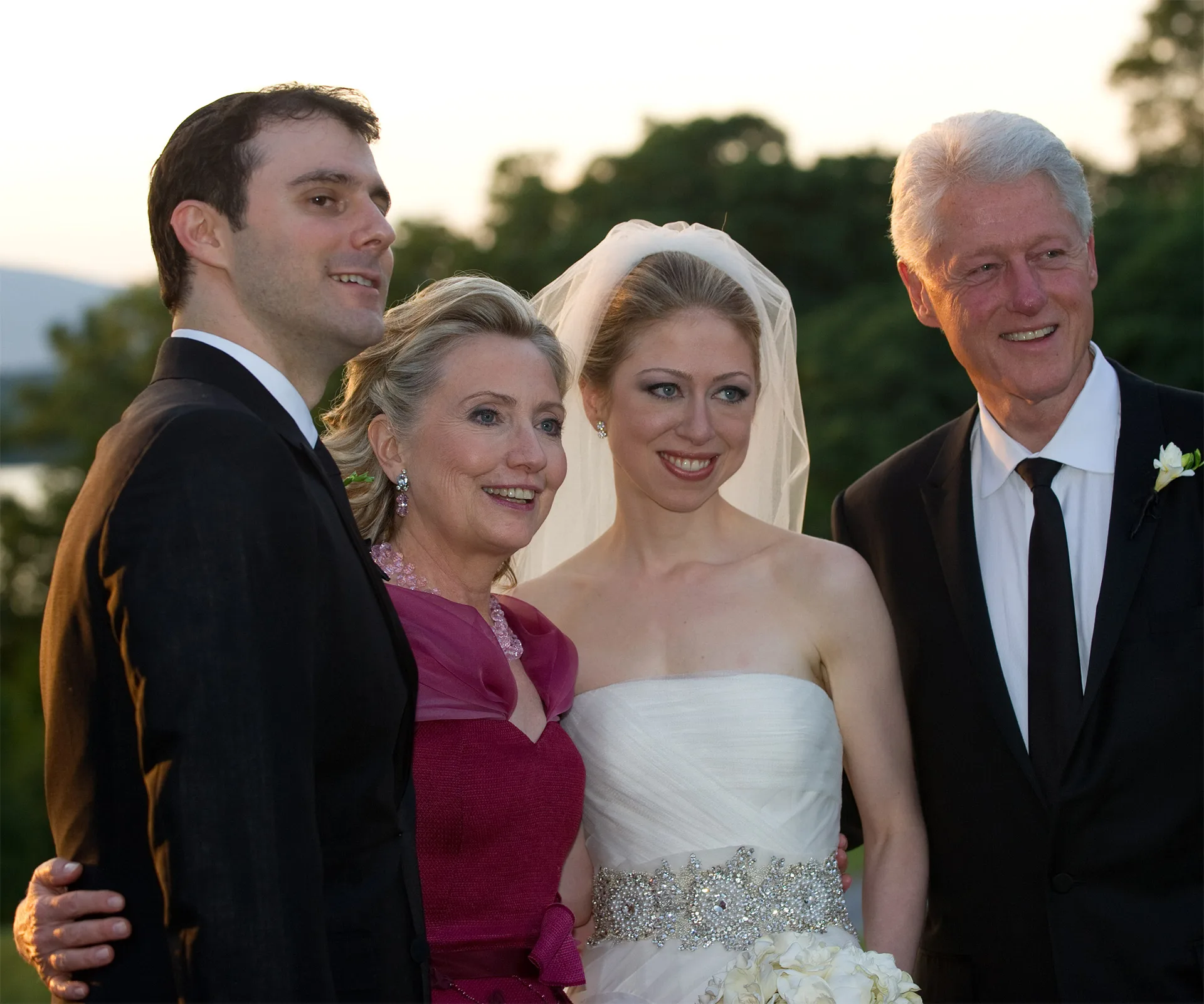 A bride and groom pose with two older people at an outdoor wedding, all smiling in formal attire.