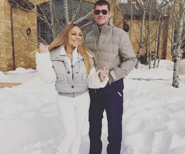 Couple posing in winter clothing, standing on snow, with a rustic building in the background.