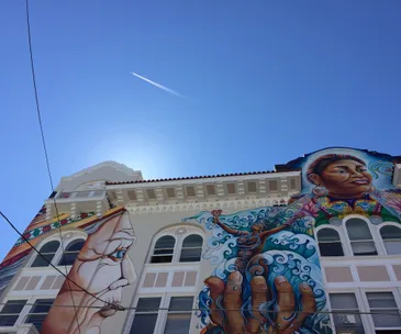 Colorful mural on a San Francisco building with faces, a hand, and abstract designs under a clear blue sky.
