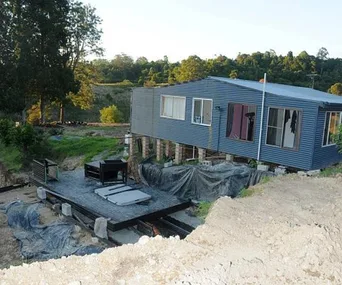 House on stilts with outdoor area under construction, surrounded by greenery and dirt excavations.