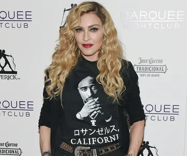 A person with long blonde hair, wearing a Tupac Shakur T-shirt, stands against a nightclub backdrop.
