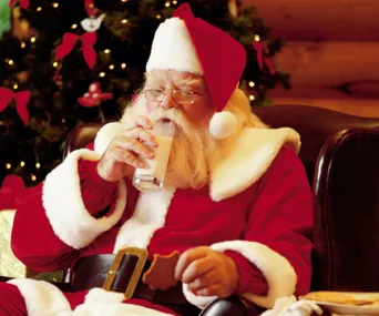 Santa Claus drinking milk and holding a cookie in a festive room with a decorated Christmas tree in the background.