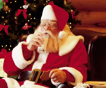Santa Claus drinking milk and holding a cookie in a festive room with a decorated Christmas tree in the background.