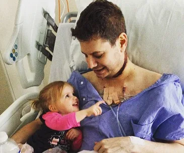 Man in hospital gown with a bandage on chest, smiling at young girl pointing at him.