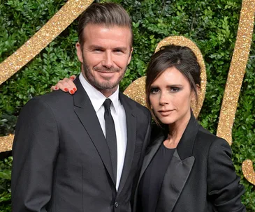 A couple in formal attire pose in front of a green foliage and gold backdrop at an event.