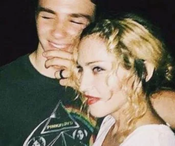 A woman with blonde curly hair embraces a smiling young man wearing a dark graphic tee.