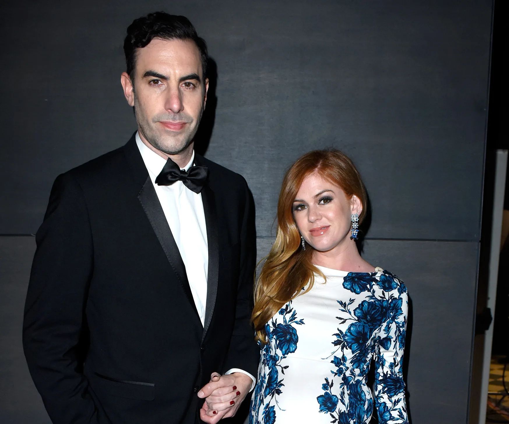 Man in tuxedo and woman in floral dress at event, standing against a dark background.