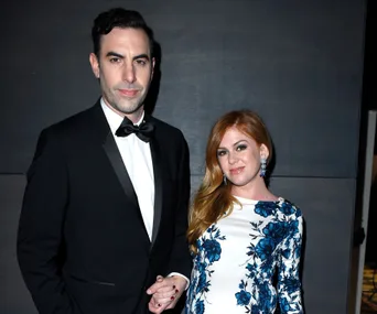 Man in tuxedo and woman in floral dress at event, standing against a dark background.