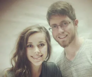 A couple smiling warmly at the camera, with the woman on the left and the man on the right wearing glasses.