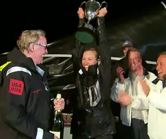 Celebration with woman holding trophy, surrounded by cheering people in wet weather gear.