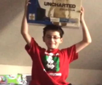 Child excitedly holds an "Uncharted Collection" PlayStation 4 box, wearing a red Mickey Mouse Christmas shirt.