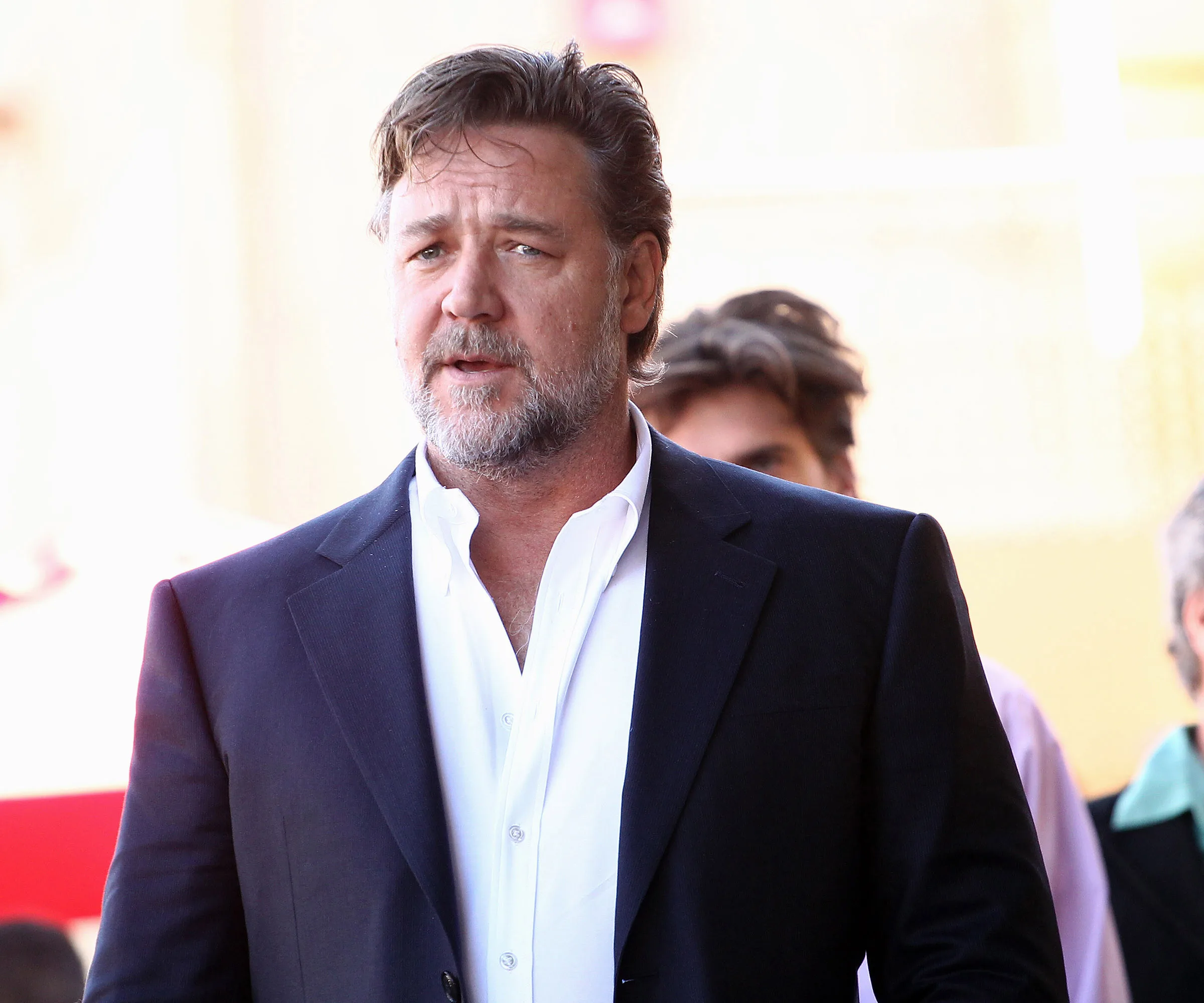 A man in a suit with a white shirt, standing outdoors, looking pensive.