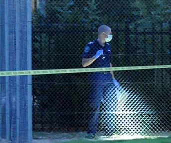 Police officer in mask investigates crime scene at night near fence, with flashlight illuminating ground.