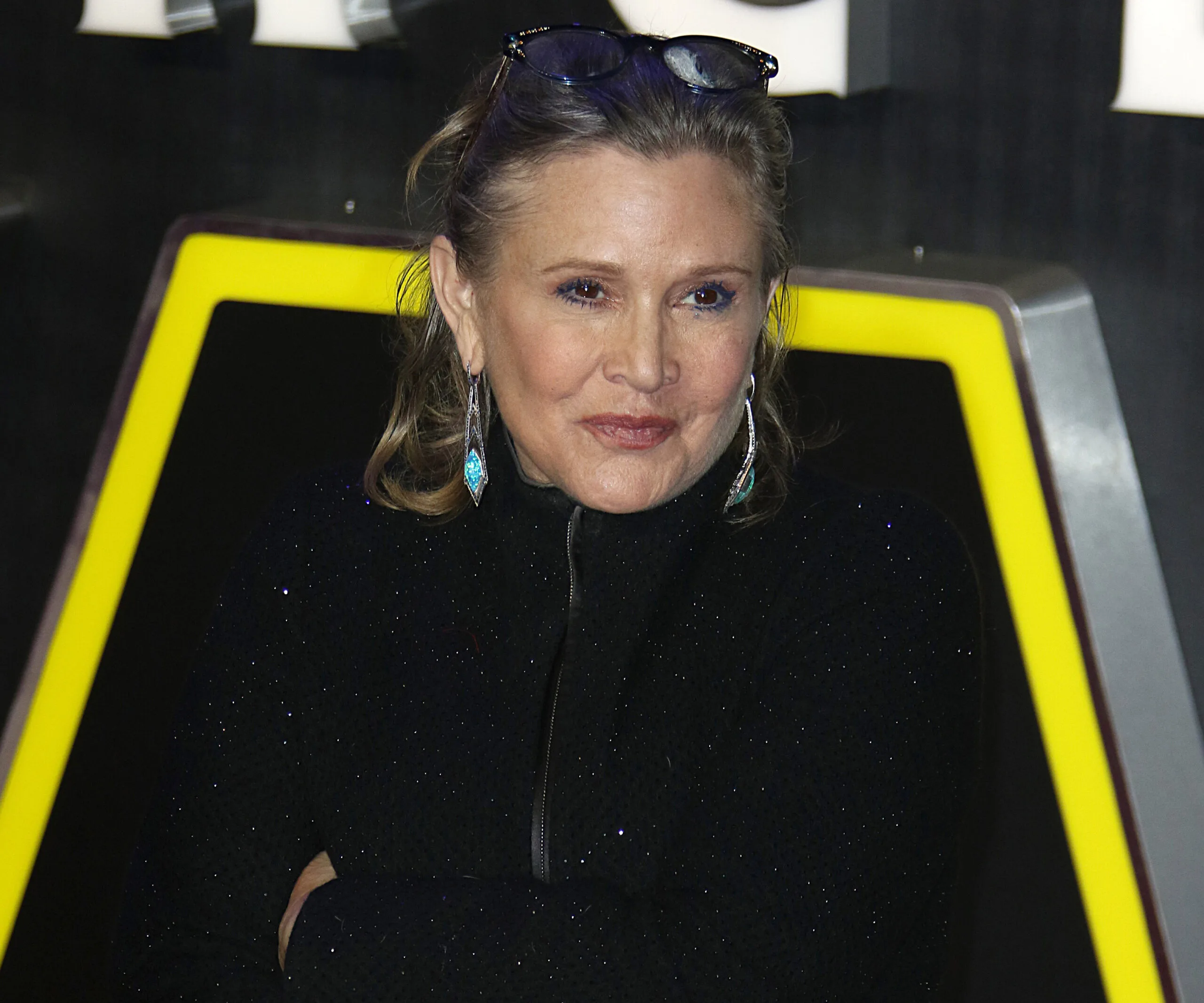 Carrie Fisher at a public event, wearing a black outfit with glasses on head and blue earrings.