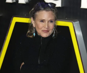 Carrie Fisher at a public event, wearing a black outfit with glasses on head and blue earrings.