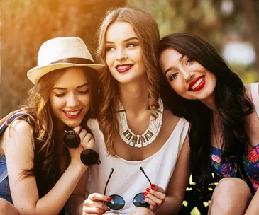 Three women smiling outdoors, one wearing a hat, another in a statement necklace, all holding sunglasses.