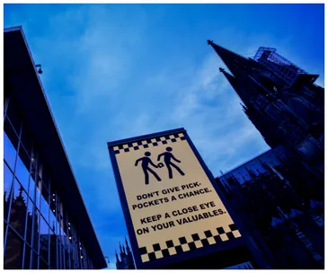 Sign reading "Don't give pickpockets a chance" near Cologne Cathedral against a blue sky.